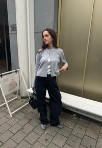 Cardigan acentuado cinza sobre uma blusa branca, combinado com calças de carga pretas. Sapatos de bico fino em preto e branco, levando uma pequena bolsa preta.