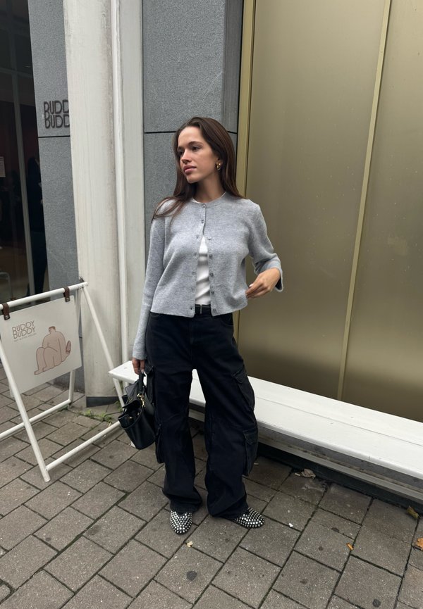 Szary kardigan z prążkowanego materiału na białej bluzce, zestawiony z czarnymi spodniami typu cargo. Czarno-białe czółenka w szpic, trzymając małą czarną torebkę.