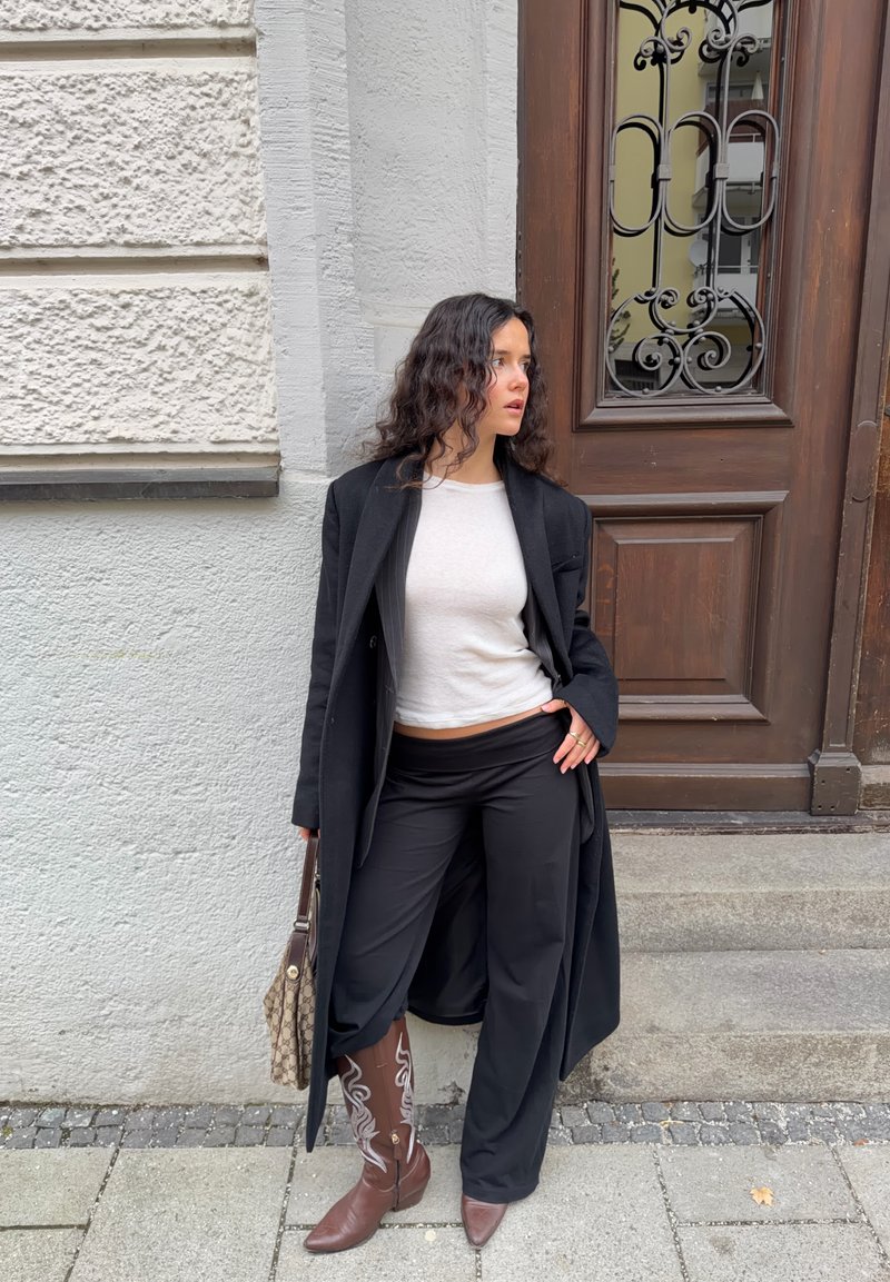 Long manteau noir sur un haut gris clair, pantalons noirs à jambes larges, et bottes de cowboy marron avec broderies blanches. Porte un sac à main à motifs.