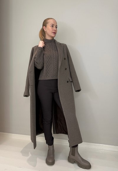 Largo abrigo de lana gris sobre un suéter texturizado gris, combinado con pantalones negros y botas de tobillo grises. El abrigo presenta doble botonadura y un diseño clásico.
