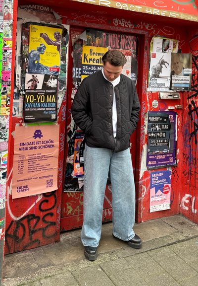 Chaqueta negra acolchada, camisa blanca, jeans holgados de color azul claro y zapatos negros, de pie frente a una pared roja cubierta de carteles y graffitis.