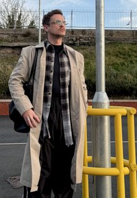 Trench-coat beige par-dessus une chemise noire et une flanelle à carreaux. Pantalon noir, bague argentée et lunettes. Debout près d'un poteau jaune. Herbe verte en arrière-plan.