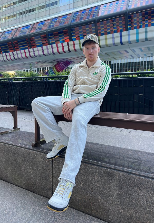 Chándal beige de Adidas con rayas verdes, pantalones de chándal gris claro y zapatillas blancas con acentos amarillos. El fondo presenta una estructura multicolor.