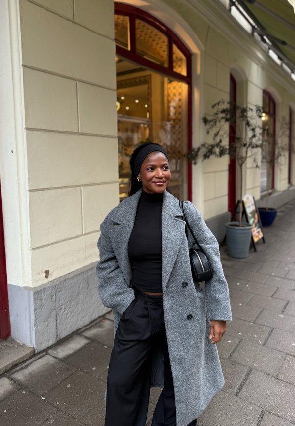 Eine Frau, die einen grauen Mantel und ein schwarzes Outfit trägt, lächelt, während sie auf einem Bürgersteig in der Stadt neben einem Gebäude mit großen Fenstern und Topfpflanzen steht.