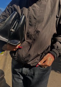 Bolsa de couro preta com um design curvado único, combinada com uma jaqueta preta mate e jeans escuros. Unhas longas e vermelhas notáveis.