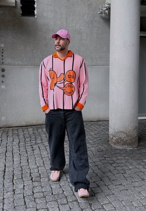 Camisa de rayas fucsias de gran tamaño con cuello y puños naranjas, que presenta un estampado gráfico juguetón. Combinada con pantalones negros holgados y zapatos rosas.