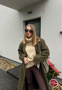 Γυναίκα με γυαλιά ηλίου, μπεζ ζιβάγκο, μακρύ χακί παλτό και τσάντα με μοτίβο λεοπάρ, στέκεται κοντά στην είσοδο ενός σπιτιού διακοσμημένη με ένα μικρό φωτισμένο χριστουγεννιάτικο δέντρο.