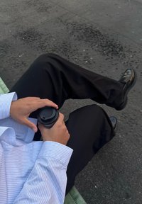 Tazza di caffè nero tenuta con due mani, vestito con una camicia a righe blu e pantaloni neri, seduto su una panchina verde con il pavimento grigio visibile.