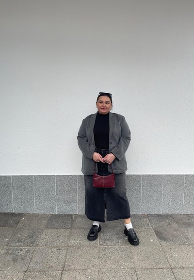 Blazer gris sobre una blusa negra, combinado con una falda de mezclilla oscura y zapatos de plataforma negros. Sosteniendo un pequeño bolso rojo. Fondo sólido.