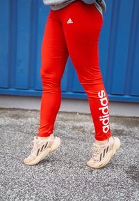 Κόκκινες αθλητικές κολάν με λευκό λογότυπο "adidas" στη αριστερή πλευρά, συνδυασμένες με μπεζ sneakers που έχουν μαύρες λεπτομέρειες και υφασμάτινες σόλες.