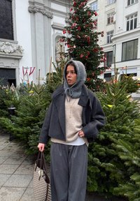 Manteau gris en laine raccourci par-dessus un pull beige, associé à un pantalon gris ample. Un foulard gris tricoté et un sac à motifs complètent le look.