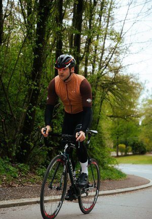 Ciclista che indossa una maglia a maniche corte arancione e marrone, leggings neri e casco, mentre pedala su una bicicletta nera con dettagli rossi su un sentiero.