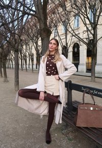 Femme en manteau beige, haut à pois et jupe ajustant des collants marron près d'arbres sans feuilles et d'un banc, tenant un grand sac à main marron en extérieur.