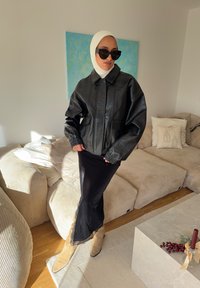 Veste en cuir noire à coupe courte, coupe ample oversize et poignets élastiques ; associée à une jupe noire élégante avec bordure en dentelle et des bottes beige.