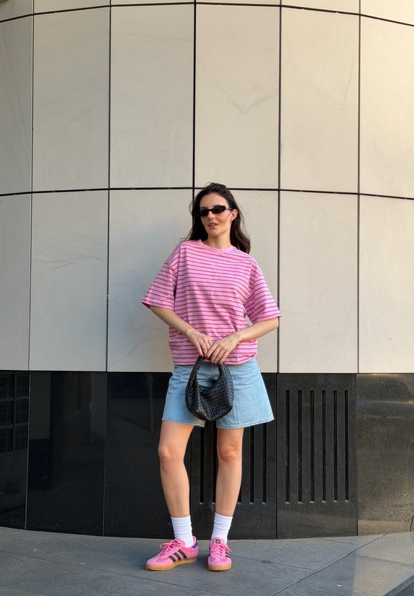 Femme en chemise rayée rose, jupe en jean et baskets roses, debout tenant un sac noir devant un mur carrelé.
