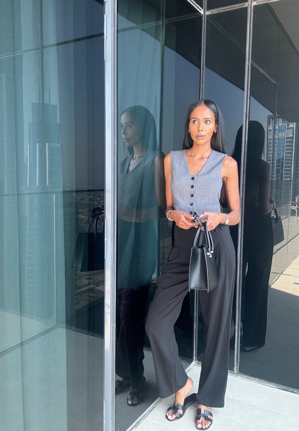 Top grigio senza maniche con bottoni, pantaloni neri a gamba larga e borsa nera. Modella in piedi davanti a un vetro riflettente con vista sulla città sullo sfondo.