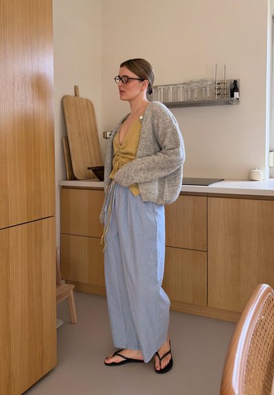 Mujer con gafas, cárdigan gris, camiseta amarilla y pantalones azul claro, de pie en una cocina de madera cerca de tablas de cortar y cristalería en una estantería.