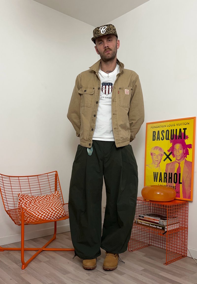 Homme portant une veste beige, un t-shirt blanc à motif graphique, un pantalon large noir et une casquette camouflage, debout dans une pièce avec une chaise en fil orange et une affiche Basquiat Warhol.