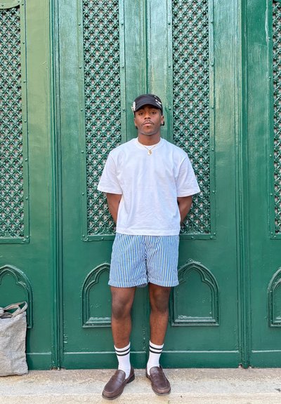 Una persona está de pie contra una pared verde con un patrón, vistiendo una camiseta blanca, pantalones cortos a rayas azules, calcetines blancos con rayas negras y mocasines marrones.