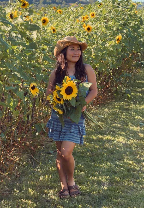 Mujer con vestido azul y sombrero de paja sosteniendo un ramo de girasoles, de pie en un campo soleado de girasoles.