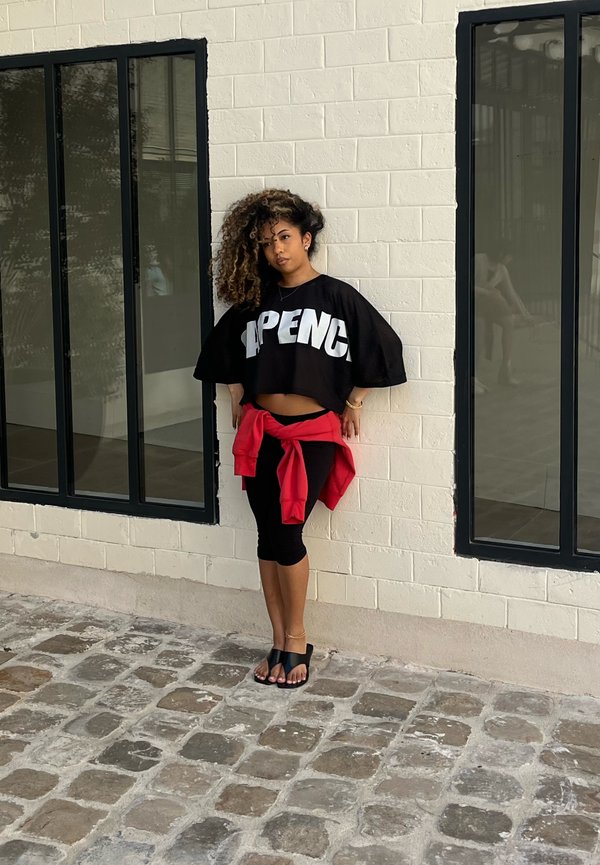 Jeune femme aux cheveux bouclés se tient contre un mur de briques blanches entre deux grandes fenêtres, portant un haut court noir et des leggings, veste rouge nouée autour de la taille.