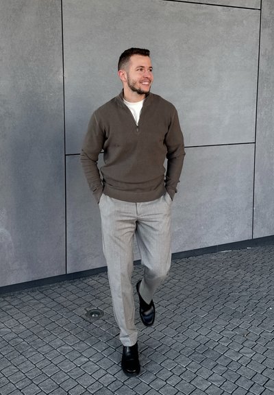 Hombre caminando sobre una acera de baldosas con las manos en los bolsillos, vestido con un suéter marrón con cremallera, pantalones gris claro y zapatos negros, sonriendo y mirando hacia la derecha.