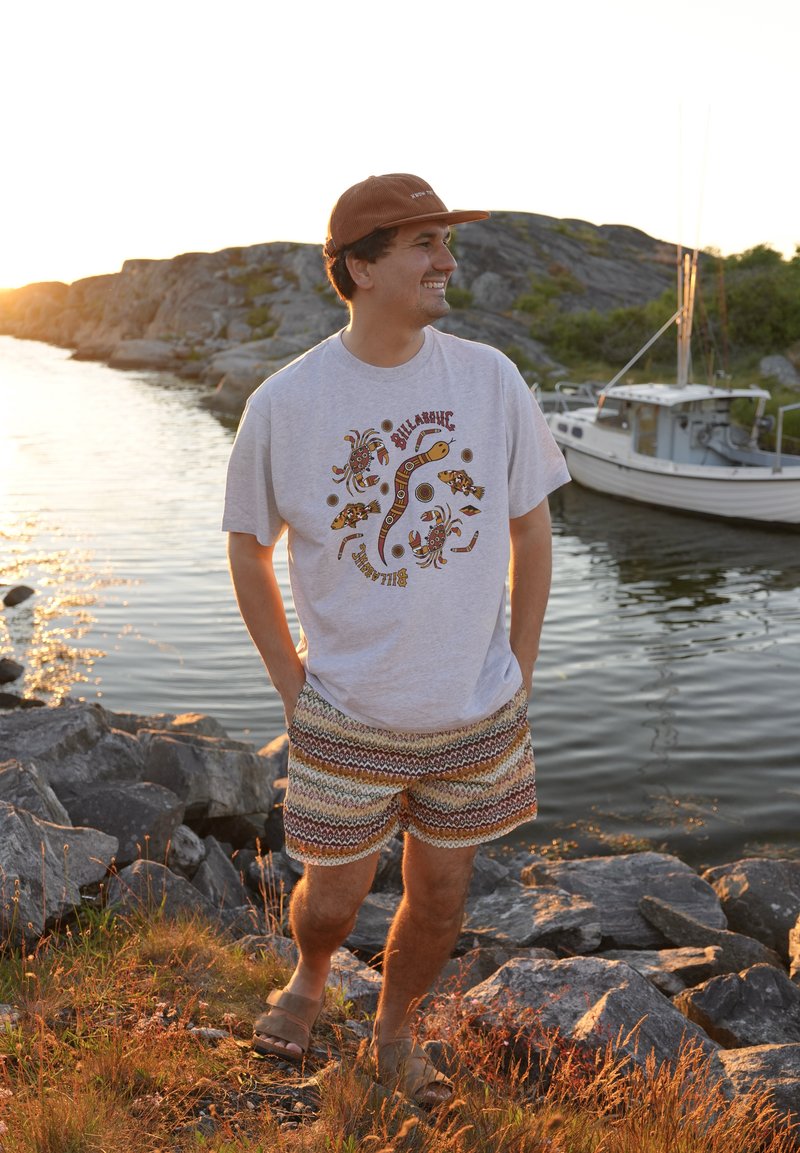 Homme souriant debout sur une rive rocheuse près de l'eau avec un bateau et des collines en arrière-plan au coucher du soleil, portant un short à motifs et un t-shirt graphique.