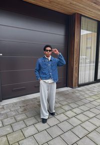 Giacca di denim con due tasche frontali, pantaloni tuta grigi e scarpe nere. Persona con occhiali da sole in piedi su un pavimento di pietra vicino a un edificio moderno.
