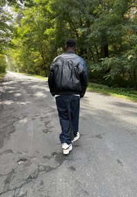 Čierna vypchávkovaná bomber bunda, voľné tmavé džínsy s dlhou šnúrkou a biele tenisky. Postava kráča po vidieckej ceste so stromami po stranách.