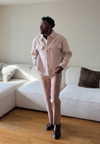Homme debout à l'intérieur portant une veste rose clair, un pantalon assorti, des chaussures foncées, avec un canapé blanc et un coussin à motif léopard en arrière-plan.