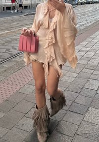 Blusa de gasa beige con volantes y cierre de lazo, pantalones cortos de mezclilla y botas de tobillo beige con flecos. Sosteniendo un bolso estructurado rosa.