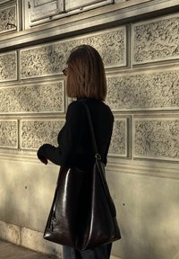 Donna con capelli alla lunghezza delle spalle, vestita di nero, che porta una grande borsa in pelle marrone scuro, in piedi contro un muro di pietra strutturata.