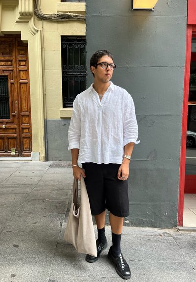 Camisa blanca de lino, pantalones cortos negros y zapatos negros. Lleva una bolsa de lona beige texturizada. De pie contra una pared gris con una puerta de madera al fondo.
