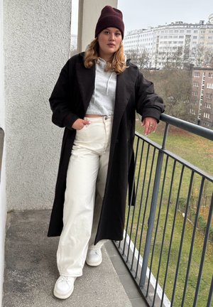 Abrigo largo negro sobre una sudadera con capucha gris recortada, combinado con pantalones blancos de pierna ancha y zapatillas blancas. La persona lleva un gorro burdeos.