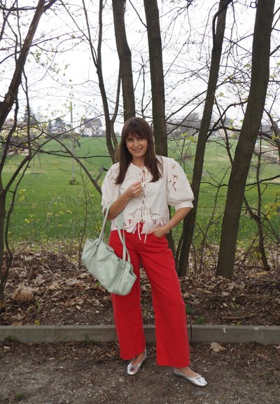 Mujer de pie al aire libre cerca de árboles, con una blusa blanca de encaje, pantalones rojos, zapatos planos plateados y sosteniendo un bolso azul claro.