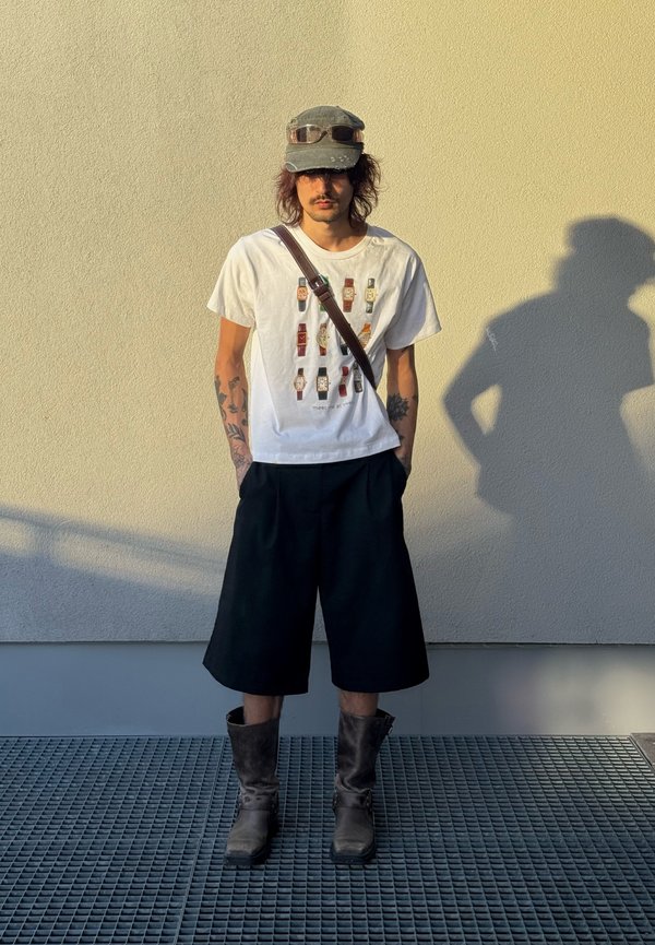 Homme tatoué portant un t-shirt blanc imprimé montre, un short noir, des bottes grises, une casquette verte avec des lunettes de soleil et un sac bandoulière, debout près d'un mur.