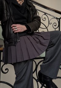 Personne portant une veste en cuir sombre, un sweat-shirt noir, une jupe plissée grise sur un pantalon gris, des bottes noires, et tenant une bague en forme d'étoile argentée à la main.