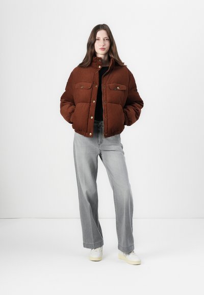 Chaqueta acolchada marrón con cuello alzado, dos bolsillos en el pecho y botones a presión dorados, combinada con pantalones vaqueros de pierna ancha gris claro y zapatillas blancas.