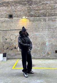 Fekete steppelt dzseki műszőrme gallérral, fekete sapka, fekete kesztyűk, túlméretezett fekete nadrág és szürke sportcipők, egy texturált fal előtt.