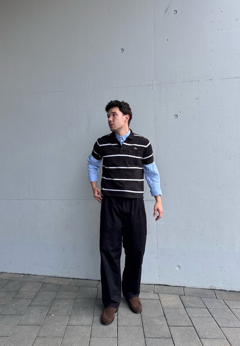 Homme portant un polo rayé noir et blanc sur une chemise bleue, pantalon noir et chaussures marron, debout sur un sol pavé devant un mur en béton gris clair.