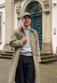 Un tânăr purtând o șapcă verde, un trench bej, un pulover gri și pantaloni negri, zâmbind în aer liber lângă o clădire cu o ușă verde și trepte de piatră.