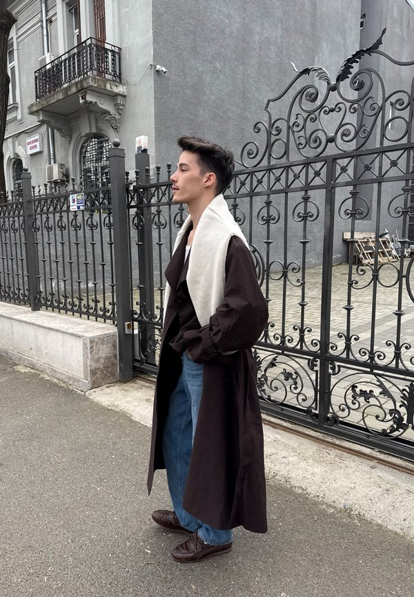 Jonge man die een lange donkere jas en een lichte sjaal draagt, staat met handen in de zakken naast een decoratief ijzeren hek en een gebouw.
