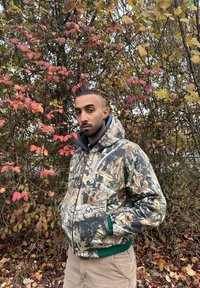 Homme portant une veste camouflage et un pantalon beige, debout les mains dans les poches devant des arbres automnaux aux feuilles rouges et jaunes.