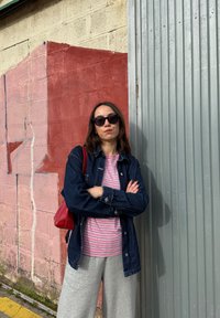 Chaqueta de mezclilla sobre una camisa de rayas rosas y blancas, pantalones deportivos grises, gafas de sol negras y un bolso rojo, frente a una pared texturizada.