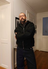 Melnā rāvējslēdzēja kapuce ar ērtu pieguļumu, vilkta pār melnu topu, kombinēta ar tumši zilām džinsu biksēm. Fona koka grīda un balti sienas.