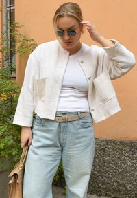 Chaqueta blanca texturizada, recortada y con botones frontales, llevada sobre una camisa blanca y unos vaqueros de cintura alta de color azul claro, acompañada de un cinturón y un bolso beige.