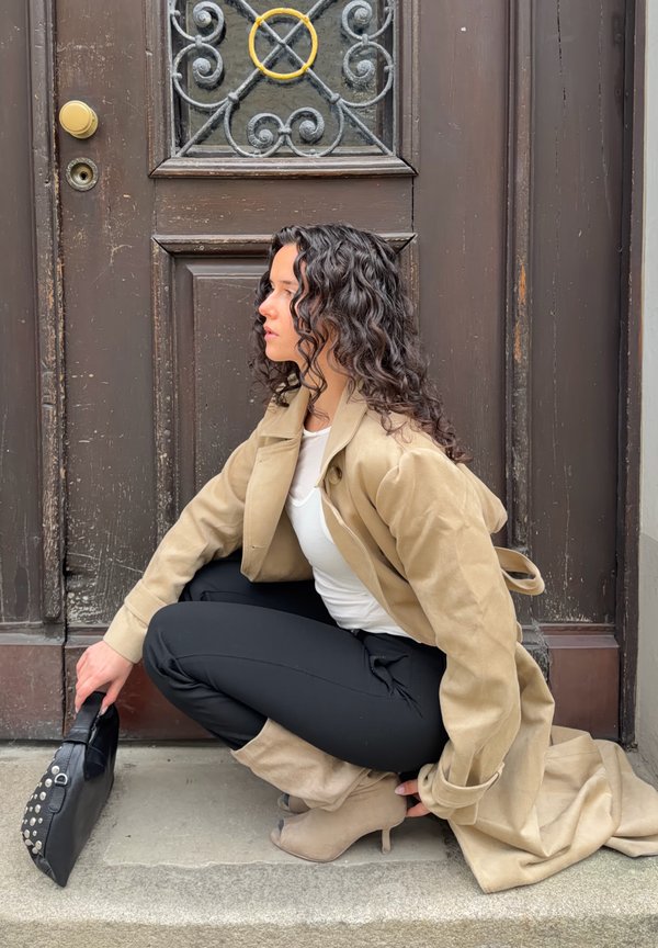 Žena s kudrnatými vlasy v béžovém trenčkotě, bílém topu, černých kalhotách a béžových botách dřepí u velkých hnědých dveří, drží černou kabelku.