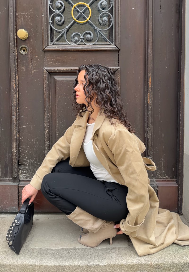 Γυναίκα με μπούκλες, με μπεζ καμπαρντίνα, λευκό τοπ, μαύρο παντελόνι και μπεζ μπότες, καθισμένη κοντά σε μια μεγάλη καφέ πόρτα, κρατώντας μια μαύρη τσάντα.