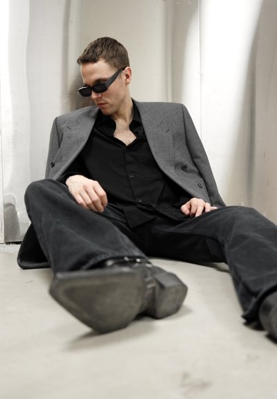 Blazer gris de doble botonadura, camisa negra abotonada y pantalones negros anchos. Gafas de sol delgadas, sentado en un suelo liso con un fondo metálico.