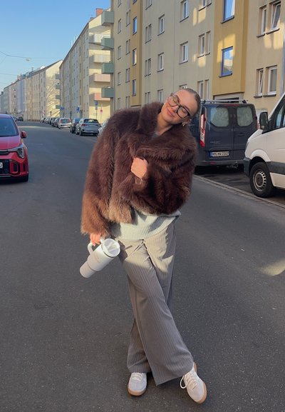 Abrigo corto de pelo sintético marrón, suéter gris, pantalones anchos a rayas, zapatillas blancas. Sosteniendo una botella de agua blanca, de pie en una calle.